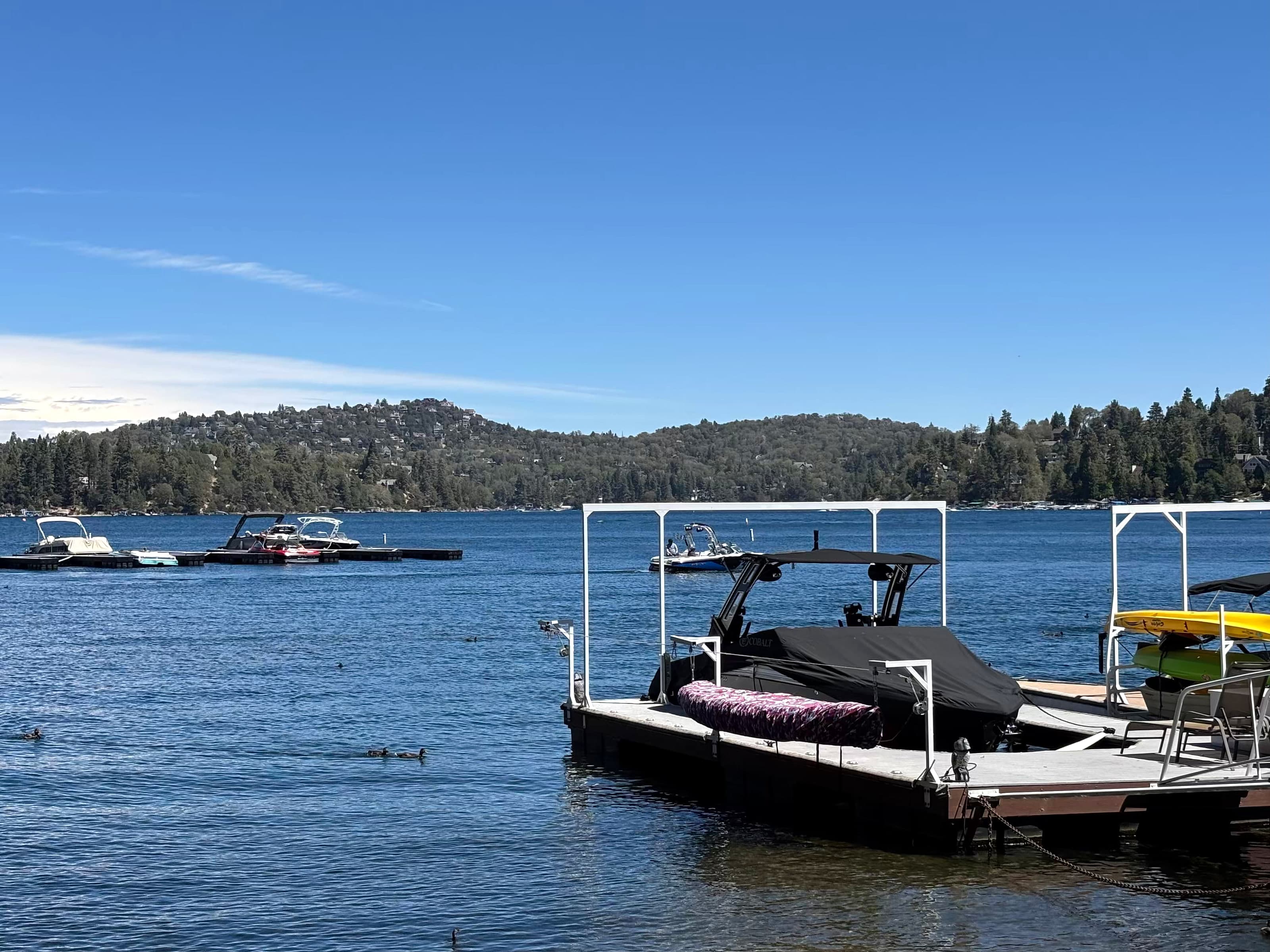 Covered motorboat and kayaks at a Lake Arrowhead dock with pine-covered mountains beyond