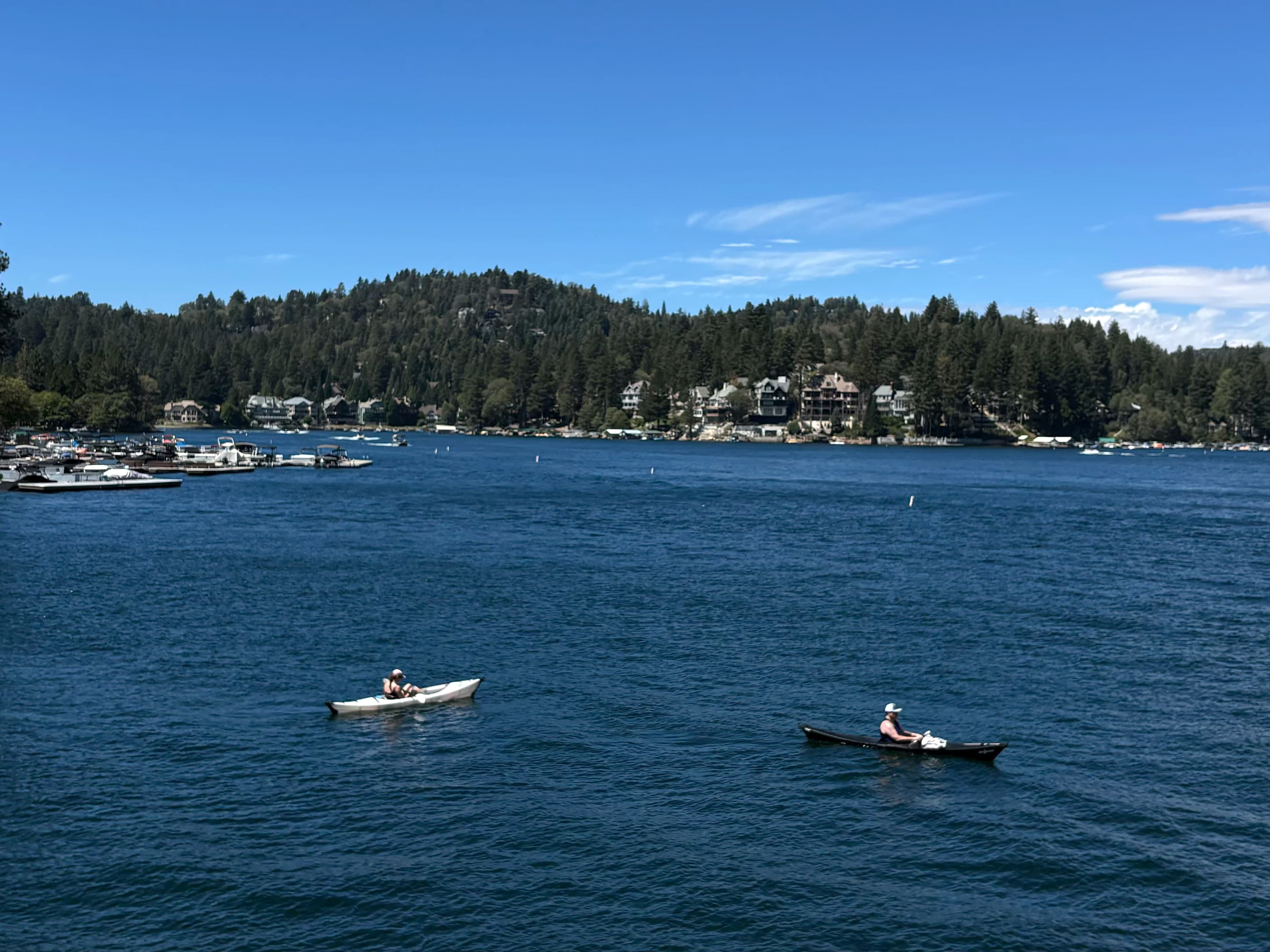 Two kayakers on Lake Arrowhead with waterfront homes along the forested shore.