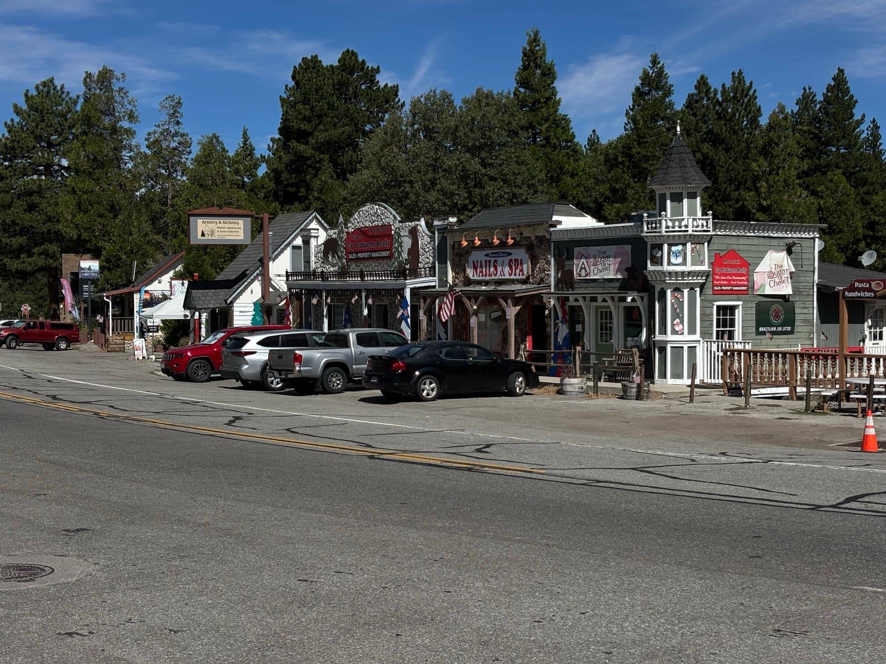 Row of mountain-town storefronts and parked cars with pine trees behind the buildings in Running Springs